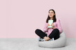 © Pixel-Shot - Young woman sitting on pouf and holding book near pink wall