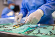 © Graphicroyalty - Close-up of a gloved hand picking up a surgical instrument from a sterile tray. Various surgical tools arranged on a green cloth in the operating room, with medical staff blurred in the background.