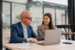 © MINAE - Senior businessman and a young businesswoman are collaborating on a project, working together on a laptop in a modern office environment, discussing strategies and analyzing data