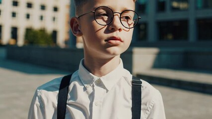 Poster - Smart young pupil wearing a white shirt, black suspenders, and round glasses stands in an urban setting, looking thoughtfully into the distance