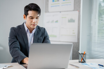 Aufkleber - Focused Asian businessman in suit is working on laptop, analyzing financial charts displayed on office wall, demonstrating dedication to his work and strategic planning