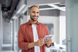 © Liubomir - A smiling, bald man in a red shirt holds a tablet in an office setting with a blurred background.