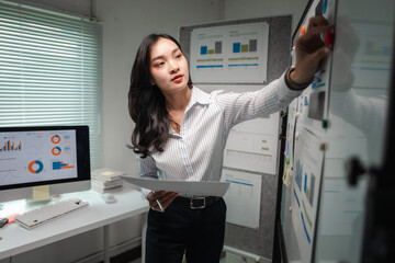 Poster - Asian businesswoman analyzing financial charts and graphs, pinning documents on whiteboard in office, working with statistics, using computer
