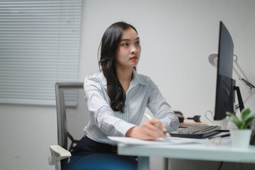 Poster - Young Asian businesswoman taking notes and working using desktop computer in office