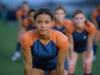 © alsu0112 - Athletic woman in blue and orange sportswear is preparing for a game on a green field, showcasing determination and teamwork among fellow players in the background