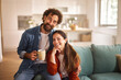 © Stockphotodirectors - A couple sits comfortably on a couch in their living room, smiling and holding a warm drink. The afternoon sunlight brightens the space, creating a welcoming atmosphere.