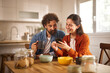 © Stockphotodirectors - A couple sits at a wooden table in their home office, smiling and sharing a moment while looking at a smartphone. They have coffee mugs and bowls of snacks in front of them.