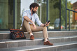 © Stockphotodirectors - A man in business attire enjoys a moment of relaxation on outdoor steps, using his smartphone while a bag rests beside him in a lively city environment.