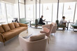 © fizkes - Comfy collaborative workspace. Open coworking space interior. Relaxing lounge zone with soft couch armchairs low table. Diverse people working on notebooks at individual desks with seats in background
