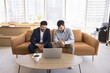 © fizkes - Building success together. 2 happy young male business partners of diverse ethnicities sit on sofa at shared office workspace create project develop startup watch video presentation on notebook screen