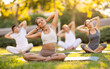 © JackF - Fit young girl enjoying group yoga session in peaceful environment of verdant summer park, performing stretches in lotus position while sitting on exercise mat