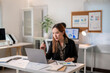 © Apichat - A woman is sitting at a desk with a laptop and a stack of papers