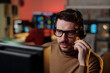 © pressmaster - Portrait of Caucasian young adult man wearing headset working at computer in scam call center, concentrating on screen with blurred monitors and coworker in background, holding microphone near mouth