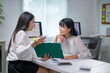 © Wasana - Two women are sitting at a desk, one of them holding a green folder