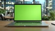 © Formoney - A sleek laptop featuring a green screen sits on a wooden desk in a contemporary office. The background showcases a vibrant urban skyline through large windows.