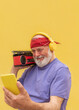 © Renata Hamuda - Smiling Senior man with headphones and boombox taking a selfie with smartphone on yellow background
