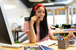 © WavebreakMediaMicro - African American woman working in open office wearing headset and writing in notebook beside laptop