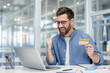 © Liubomir - A man celebrates a successful online purchase, holding a credit card near a laptop in an office setting. He is joyful.