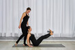© TRAVELARIUM - Two young adult women in black athletic wear engage in yoga practice. One, seated on mat, elevates her legs with support from block, assisted by other, creating calm, focused atmosphere