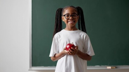 Poster - Happy african american schoolgirl wearing white t shirt and glasses is holding a red apple while standing in a classroom in front of green chalkboard