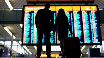 Poster - Businessman and businesswoman holding hands and looking at timetable of flights, checking their flight on the information board at the airport