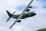 C-130 Hercules military transport aircraft is captured in flight from below, with its four propellers visible against a cloudy sky