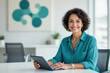 © f_bossa - A smiling woman with curly hair sits at a desk, holding a tablet and looking directly at the camera in a modern office setting.