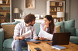 © Stockphotodirectors - In a cozy living room, a couple engages in a serious conversation about their financial issues while examining documents and utilizing a laptop for information.
