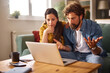 © Stockphotodirectors - A couple appears stressed as they sit on a couch, looking at a laptop. They are surrounded by documents and a coffee mug, indicating a serious discussion about financial issues.