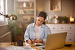 © Stockphotodirectors - A woman sits at a wooden desk in a cozy home office, writing in a notebook with a laptop open nearby. Natural light fills the room, creating a warm and productive atmosphere.