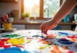 © starush - Child's hand dips brush into colorful paint on a creative art table in a bright kitchen