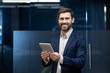 © Tetiana - Portrait of a smiling and successful young businessman in a suit standing in an office, holding a tablet and looking at the camera