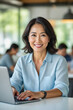 © f_bossa - A smiling Asian woman in a light blue shirt sits at a desk, typing on a laptop in a modern office setting.