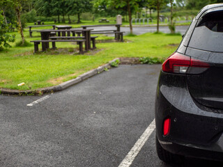 Wall Mural - Black family car parked in a parking lot of a park with barbecue or dining area, selective focus. Nobody. Car surface is with rain drops. Outdoor activity. Travel and tourism.