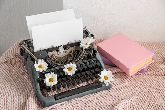 Vintage typewriter with paper sheet, books and daisies on knitted blanket near white wall