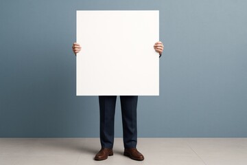 Poster - Businessman in formal attire holding large blank white square mockup against blue wall background for advertising or branding concept.