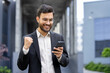 © Liubomir - A young businessman celebrates a success while looking at his phone. He's wearing a suit and glasses, with an elated expression.