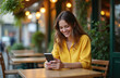 © Vadym - Young woman smiles joyfully using smartphone at cafe. Browsing internet on mobile device. Happy female in yellow shirt is chatting, texting, reading, or working outside. Internet lifestyle.