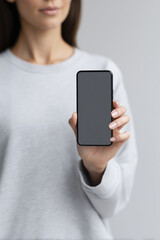 Fototapete - Young woman in gray sweatshirt holding smartphone with blank screen, showcasing modern technology and digital communication, perfect for mockup and design purposes