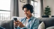 © Natsumae - Smiling young man sits comfortably on a sofa, wearing headphones and using his smartphone, enjoying his music in a relaxed home setting.