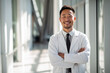 © MobiWall - Asian American male doctor in white lab coat with black tie, smiling confidently with arms crossed while standing in hospital hallway. Modern healthcare and professionalism concept.