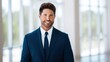 © na - Confident businessman in a dark blue suit and tie, smiling directly with a well-groomed beard, styled brown hair, indoors with bright windows and blurred greenery.