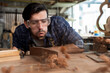 © jittawit21 - Carpenter blowing sawdust off wood surface while working in workshop, wearing safety goggles, close-up