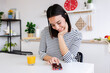 © Xavier Lorenzo - Cheerful pretty young woman using her smart mobile phone and enjoying orange juice sitting in a cozy kitchen at home, enjoying social media content in the morning