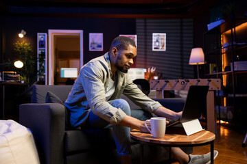 Poster - Adult guy typing on laptop laying on his couch at his stylish apartment, engaging in freelancing and online work. Black person multitasking efficiently and embracing the flexibility of remote job.