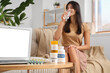 © Pixel-Shot - Young woman taking pills at home, focus on bottles with different vitamins B and laptop