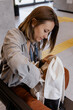 © MarijaBazarova - Young woman sitting in a modern waiting area and searching inside her white backpack, concept of travel, everyday routine, and preparation for a journey