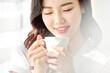 © Dialogue Frame - Young Asian Female Standing by Window Holding Coffee Cup in Bright Morning Light