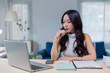© Tj - Young woman working at a home office, focused on her laptop. She is surrounded by notebooks and a smartphone, indicating a productive and organized workspace
