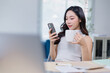 © Tj - Young professional woman multitasking at her desk, holding a coffee mug and using a smartphone. Bright office setting with natural light and modern decor in the background